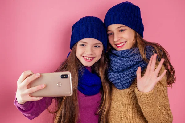 Twee mooie tienervriendinnen knuffelen de winter met gebreide hoeden die selfie nemen op de smartphone. jonge vrouwelijke vrienden zussen portret in studio roze achtergrond.herfst winter mode look seizoensgebonden verkoop — Stockfoto