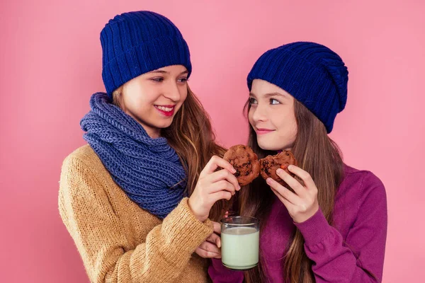 아름다운 10 대 여자 친구 둘 이 겨울을 나면서 모자를 쓰고 쿠키와 뜨거운 우유를 마시고 있습니다. 젊은 여자 친구들의 사진 이 스튜디오 핑크 백 그라운드에 있다. 크리스마스 밤, 잘 자려고 술을 마신다. — 스톡 사진