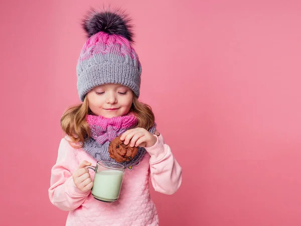 Güzel küçük kız sütü içer ve örgü bir şapka ve eşarpla kurabiye yer. Stüdyoda, Noel gecesi, pembe bir arka planda. — Stok fotoğraf