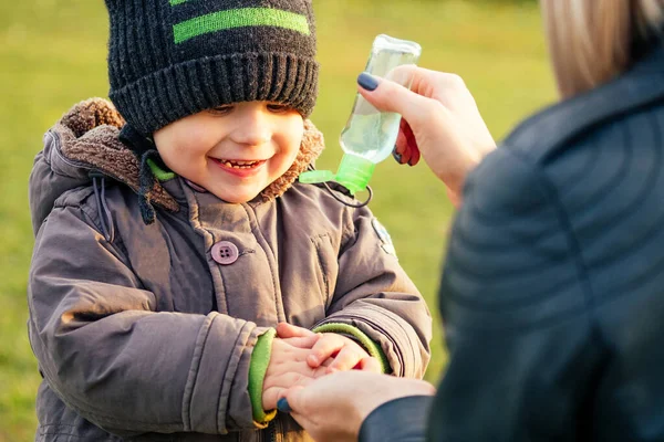 Mutter in warmer Jacke und Strickmütze trägt ein antiseptisches Gel auf, antibakterielles Gel. stylische blonde Frau mit Kind vor der Kulisse des Sees — Stockfoto