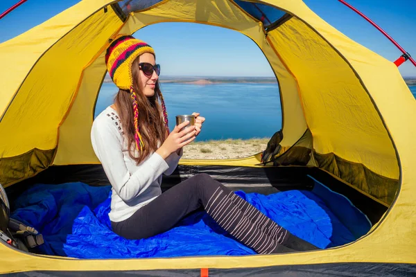 Jovem mulher em um chapéu engraçado do Nepal sentado em um saco de dormir azul na tenda segurando uma caneca térmica e beber xícara de café chá fundo paisagem lago. bom dia na viagem — Fotografia de Stock