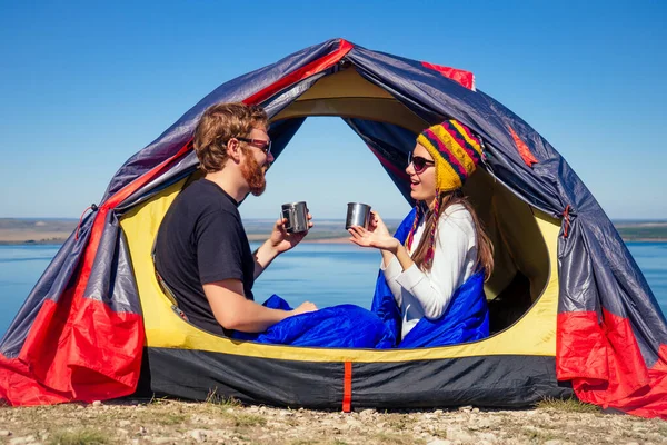 Casal de viagem no amor acampar em rocha com vista para o lago. Jovem mulher e homem feliz pessoas sentadas na tenda envolto em um saco de dormir segurando uma caneca de xícara de café de chá. bom dia ritual — Fotografia de Stock