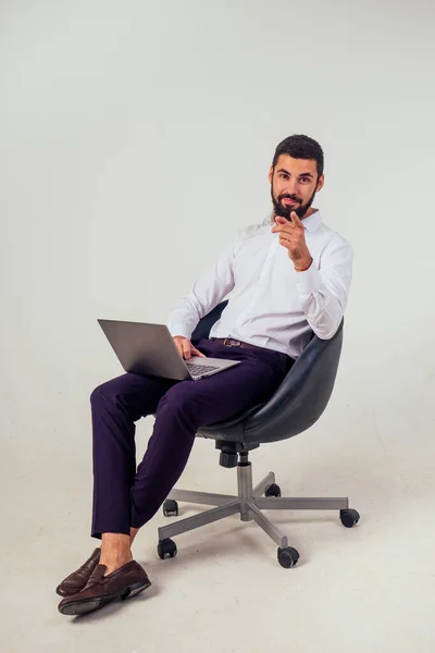 Trabajador de oficina masculino seguro y guapo sonriendo y sosteniendo un ordenador portátil sentado en una silla sobre un fondo blanco en el retrato de larga duración del estudio — Foto de Stock
