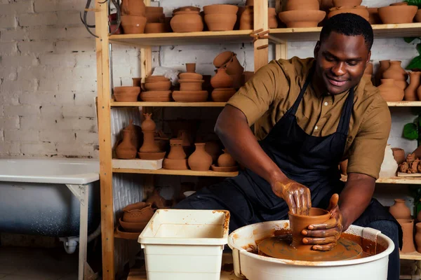 Siyah önlüklü ve şık koyu tişörtlü melez afro erkek çömlekçi atölye masası çarkında oturuyor, el yapımı kil kahverengi vazoya jöle sürüyor.