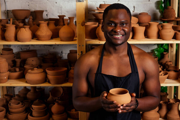 young and confident afro american man in a black apron sculpts a potter from a clay pottery workshop. clay pot in the hand of the latin artist
