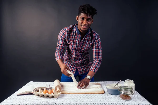 Yakışıklı ve genç afro Afrikalı adam ev yapımı pastalar hazırlıyor. Un ile kirlenmiş taze hamur işlerinden Amerikan pastası. Masada yumurta, atölyede siyah bir arka planda merdane ve yemek tarifi kitabı var. — Stok fotoğraf