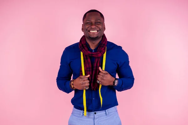 Homem afro-americano trabalhando em uma oficina de costureira alfaiate estiloso modelo masculino designer de roupas com uma fita métrica no pescoço posando em um fundo rosa no estúdio — Fotografia de Stock