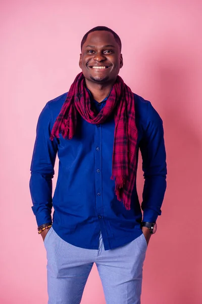 Bonito jovem africano de camisa azul, calça cinza e um sorriso de cachecol vermelho em um fundo rosa na oficina do estúdio — Fotografia de Stock
