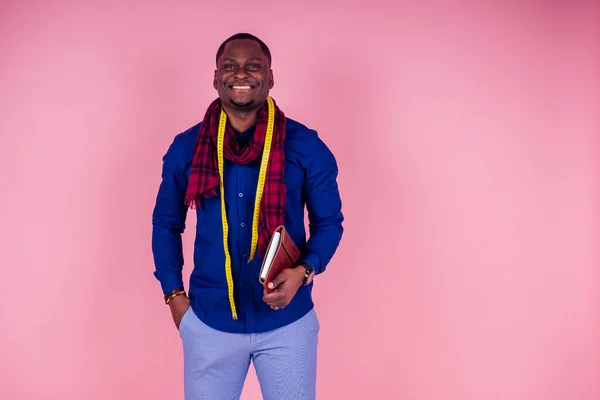 Homem afro-americano trabalhando em uma oficina de costureira alfaiate estiloso modelo masculino designer de roupas com uma fita métrica no pescoço posando em um fundo rosa no estúdio — Fotografia de Stock