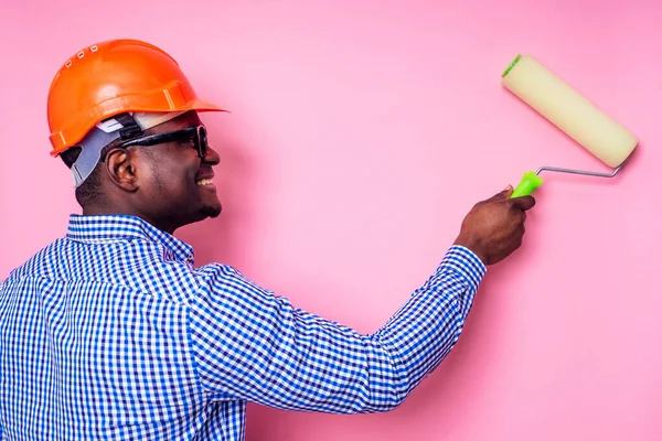 Black man African American holding paint roller in hand paints the wall in pink color .happy african builder painting inside the house,businessman wears a helmet hard hat.young guy is painting — Stock Photo, Image