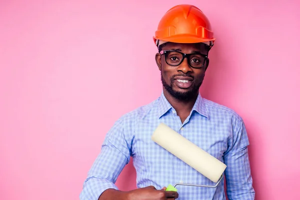 Černý muž Afroameričan drží barvu váleček v ruce maluje stěnu v růžové barvě.šťastný africký stavitel malování uvnitř domu, podnikatel nosí helmu tvrdý hat.young chlap je malování — Stock fotografie