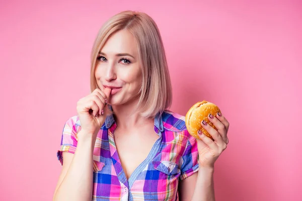 Blondine perfekte Haut essen Burger. das schöne Mädchen isst appetitlich einen Hamburger. ein Student mit einem vegetarischen Sandwich auf rosa Hintergrund — Stockfoto