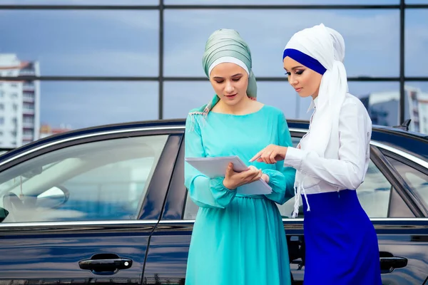 Duas mulheres empresárias muçulmanas europeias bonitas e bem sucedidas no elegante hijab e turbante segurando papel arranha-céu escritório janelas de fundo na rua de pé perto de seu carro preto — Fotografia de Stock