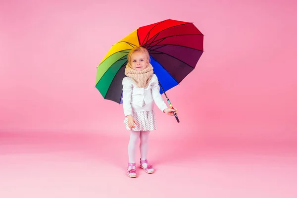 Portrait of happy and beautiful little blonde girl blue eyes in beige knitted scarf holding a colorful rainbow umbrella autumnal springtime season in the studio on a pink background.charming child — Stok fotoğraf