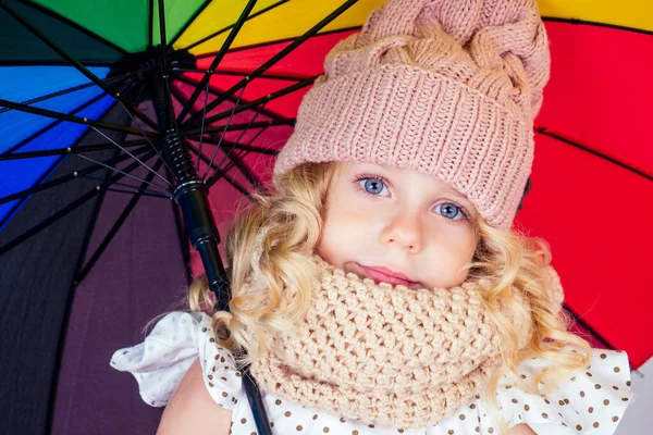 Renkli bir gökkuşağı şemsiyesi taşıyan bej örülü pembe şapkalı mutlu ve güzel sarışın kız portresi. İlkbahar mevsimi. Parkta yürüyen büyüleyici çocuk. — Stok fotoğraf