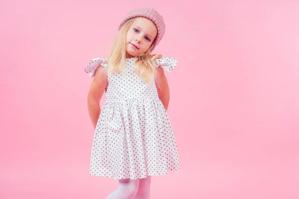 Body portrait of adorable little girl blonde girl blue eyes model in beige knitted pink hat in white princess dress in peas autumnal springtime season in studio shot on pink background — Stock Photo, Image