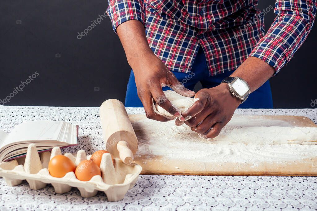 African American male cook chef boasts of pastry hands in flour baking .man s hands sheets dough with rolling pin bakes cake , the eggs and recipe book on the table. homemade baking