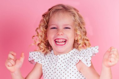 Küçük kız, mavi göz, sarı saç stili, başında taç, üzerinde bezelye desenli şirin beyaz bir elbise stüdyoda pembe bir arkaplanda poz veriyor. Doğum günü kutlaması, güzellik yarışması.