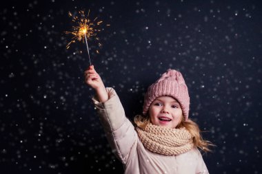 charming little girl in a knitted pink hat holding fireworks on black background in a studio.Cute blonde child with xmas dream.Happy kid enjoy the fire sparks. new year holidays eve of Christmas wish clipart