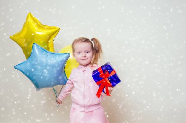 Küçük şirin kız elinde helyum mavisi, sarı ve altın renkli balonlar ve beyaz arka planda bir hediye kutusu tutuyor. Noel ve yeni yıl tatili.