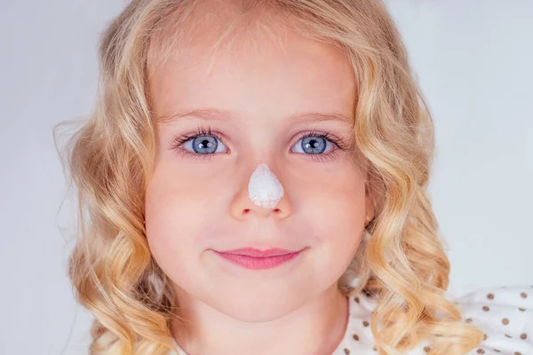 Little and beautiful blonde curly hairstyle girl in cute dress blot sunscreen on the face. blue eyes, perfect skin, on the nose spf cream close up portrait on white background in studio — Stock Photo, Image