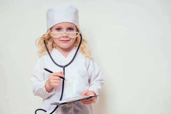 Uśmiechnięta dziewczynka w mundurze lekarza z narzędziami medycznymi stetoskop pisząc coś do schowka na białym tle w studio kopiuj space.future zawód poradnictwo zawodowe kariera — Zdjęcie stockowe