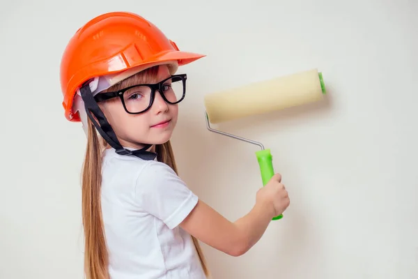 cute and little girl in a construction helmet hard hat and glasses holding paint roller paints a white wall at home.future designer business woman architect career guidance concept