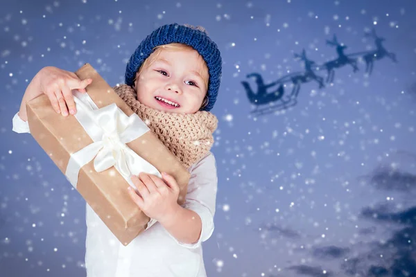 Portrait de petit garçon avec un cadeau dans un chapeau tricoté sur fond bleu foncé.Mignon enfant blond avec cadeau de Noël. Heureux gamin profiter des vacances. Père Noël volant en traîneau la veille de Noël ciel neigeux — Photo