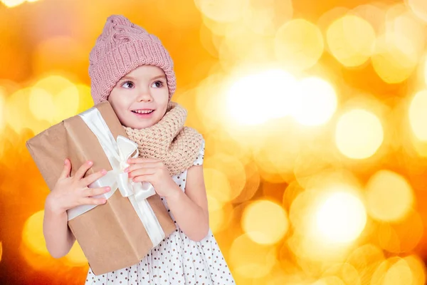 Close-up portrait of a little happy and beautiful blonde girl in a dress, hat and knitted scarf holding in hand box with a new year gift with white bow bright yellow garland sparks blurred background — 스톡 사진