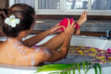 Küvette köpükle ve tropik çiçeklerle rahatlayan genç bir kadın, eko otel Bali.brunette 'de tropikal çiçek yaprakları bedeni jakuzide süngerle sabunluyor cennetin kumsalında egzotik bir yerde.