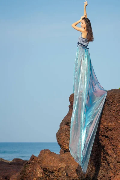 Розкішна брюнетка молода жінка в синій сукні хамелеона довгий потяг, що стоїть на скелі біля моря, пісок і тропічні дерева, великі хвилі б'ються на скелі.красива русалка самотня меланхолія чекає — стокове фото