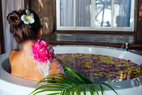 Jovem mulher bonita relaxando no banho com espuma e pétalas de flores tropicais — Fotografia de Stock