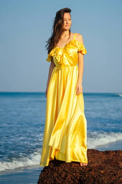 Красива жінка з літаючим довгим метеликом шовково-жовта сукня позує на пляжі, стоячи на скелі — стокове фото