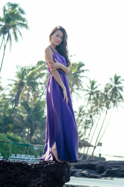Красива молода жінка в стильній фіолетовій довгій шовковій сукні, насолоджуючись день на пляжі — стокове фото