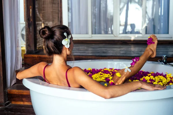Mulher com plumeria no cabelo em sutiã vermelho relaxante em banho redondo com flores tropicais, cuidados com a pele orgânicos, luxo spa eco hotel — Fotografia de Stock