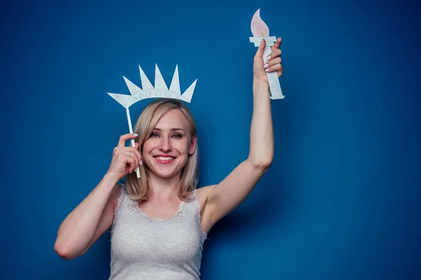 Mujer rubia sosteniendo bandera americana con corona de papel y antorcha Estatua de la libertad sobre un fondo azul en el estudio. 4 de julio concepto de celebración del día de la independencia y aprendizaje del idioma inglés — Foto de Stock