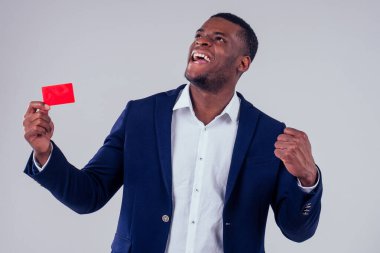 Siyah pantolonlu ve ceketli şık bir Afro-Amerikan. Stüdyoda beyaz arka planda kırmızı kredi kartı tutan büyük bir gömlek yakalı.
