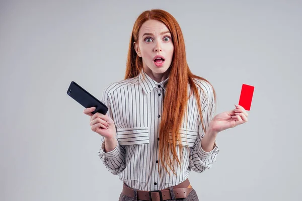 Рыжеволосая деловая женщина в полосатой рубашке счастливая с помощью мобильного телефона и держа красный картон пластиковой карты на белом фоне студии черная пятница распродажа — стоковое фото