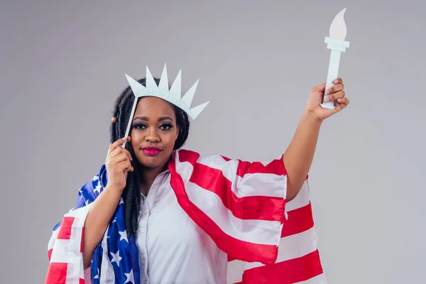 Afroamerikanische Armee Latein in einem weißen medizinischen Kleid Kleidung Dreadlocks Make-up und große Lippen mit einer Papierkrone und einer Fackel auf weißem Hintergrund im Studio mit amerikanischer Flagge, Unabhängigkeitstag USA 4. Juli — Stockfoto