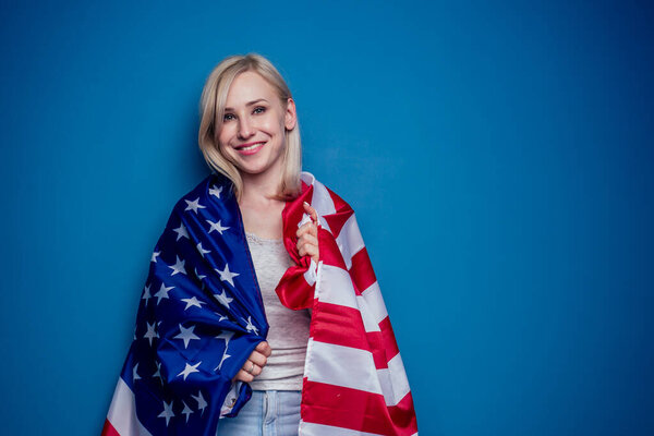 blonde woman in jeans holding American flag with paper crown and torch Statue of liberty on a blue background in the studio .4th july independence day celebration concept