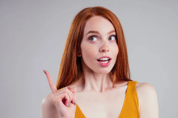 Счастливая весёлая рыжая молодая женщина с идеальной кожей в жёлтой рубашке, смотрящая вверх и указывая пальцем на подражание, думает о белом студийном фоне. Концепция идеи и вдохновения — стоковое фото