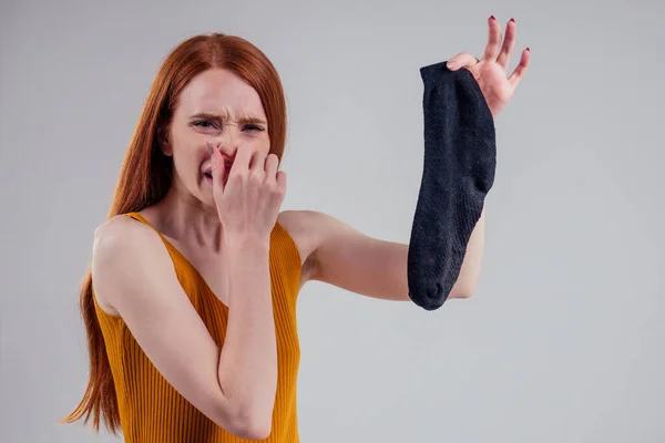 Pelirroja hembra en un gesto de camisa amarilla huele mal sosteniendo un calcetín negro sucio hacia fuera una mirada asquerosa en su cara en el estudio fondo blanco . — Foto de Stock
