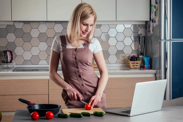 Una bella donna bionda che utilizza il computer portatile in cucina e tagliare le verdure per la cottura dell'insalata e guardare un film, ricetta online videoconferenza chat — Foto Stock
