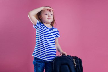 Büyük bir seyahat bavulunda oturan çizgili mavi tişörtlü kadın çocuk rüya sinekleri deniz kıyısındaki pembe arka plan stüdyosunda dinleniyor.