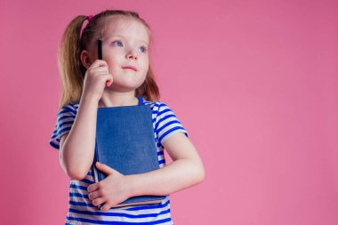 Denizcilik tarzı seyahat tarzı bavul giyen küçük bir kız deniz kıyısındaki pembe arka plan stüdyosunda dinleniyor ve elinde bir ders kitabı tutuyor.