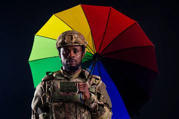 Ejército militar africano guerrero traje de camuflaje tristeza envuelto en una bandera americana de pie bajo un paraguas de arco iris estudio de fondo negro, mentir noticias violencia medios criminales — Foto de Stock