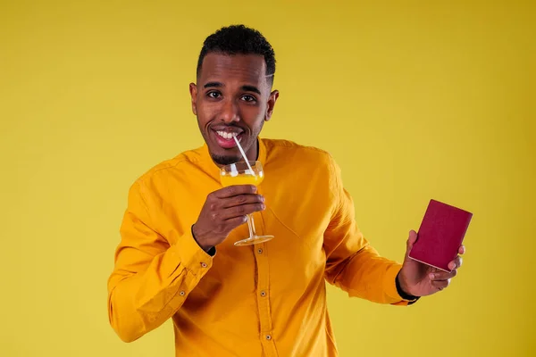 Bonito feliz jovem africano homem Quênia África turista isolado amarelo parede fundo segurando bilhetes com passaporte e coquetel viagem de vidro ao redor do mundo — Fotografia de Stock