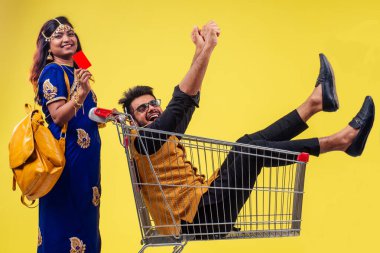 Hintli güzel kadın, stüdyo sarı arka planda alışveriş yapan metal kamyonda mutlu bir erkek taşıyor. Kara Cuma satış sezonu konsepti