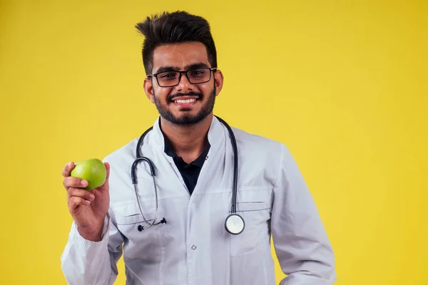 Indian male in medical uniform eating apples in yellow studio background — Stock fotografie