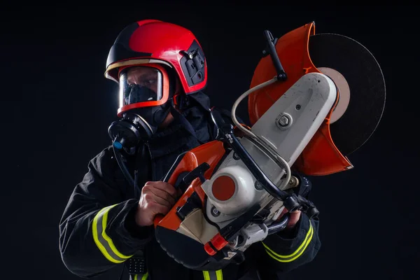 Retrato forte bombeiro em uniforme à prova de fogo segurando um machado motosserra em suas mãos preto fundo estúdio — Fotografia de Stock